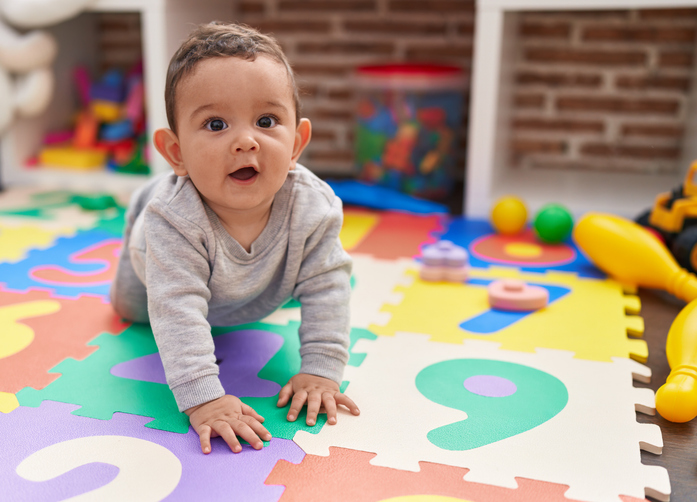 Happy baby boy crawling on a colorful foam alphabet play mat with toys scattered in the background.