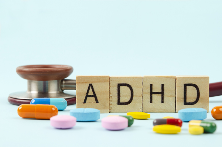 Wooden blocks spelling ADHD next to a medical stethoscope and scattered colorful medication pills on a light blue background.