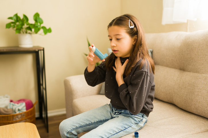 Young girl holding her throat while preparing to use a blue asthma inhaler on a couch at home.