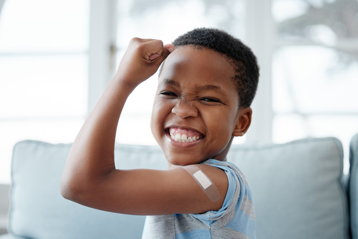 Smiling young boy flexing his arm with a adhesive bandage on his shoulder after getting a vaccine injection.