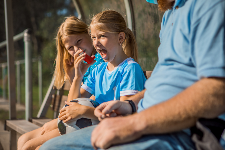 Young soccer player sitting on the bench using a red inhaler during an asthma attack.