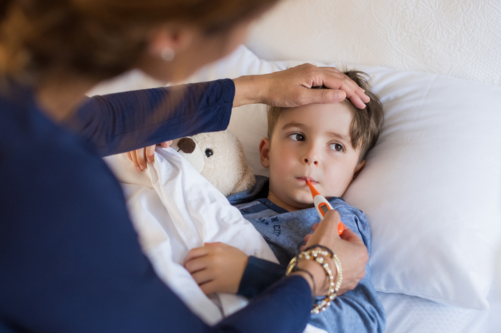 Mother checking her sick young boy's temperature with an oral digital thermometer while he lies in bed with a teddy bear.