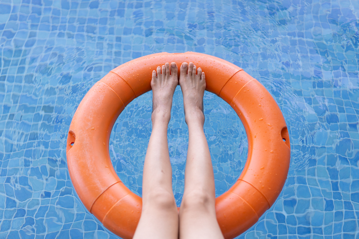 Person resting their legs on an orange lifebuoy ring floating in a blue tiled swimming pool.