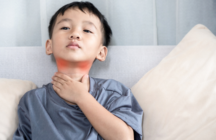 Young boy sitting on a couch holding his throat in pain, with a red highlighted area indicating a sore throat.