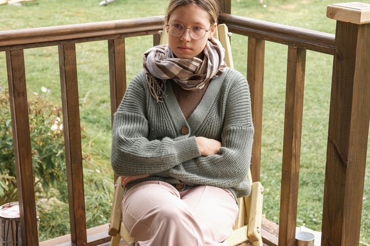 Sad teenage girl with glasses sitting on a porch chair with arms crossed, looking away thoughtfully.