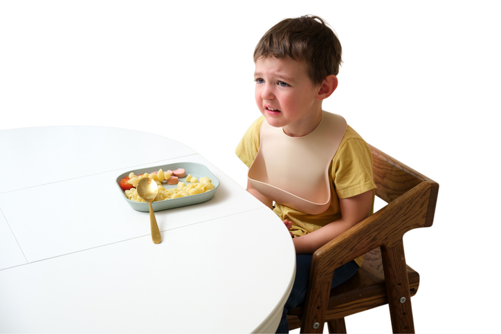 Crying toddler boy wearing a bib at a white table, refusing to eat a plate of pasta and sausage.