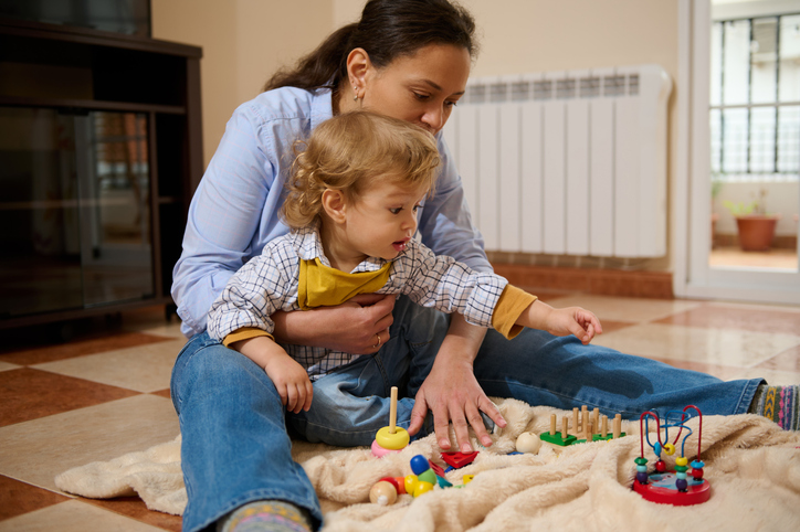 Mother supervising her toddler boy as he reaches for colorful wooden educational toys on the living room floor.