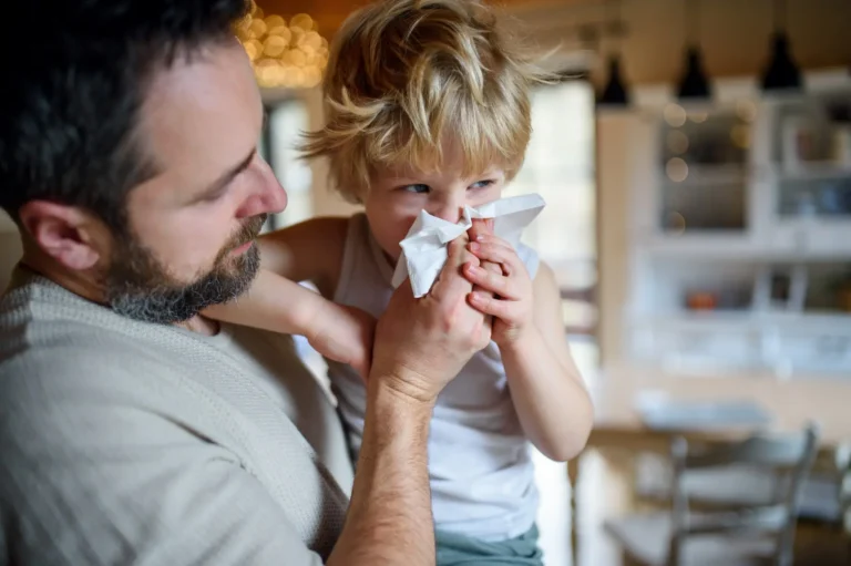 Parent holding a child with a runny nose as the child uses a tissue, illustrating common cold, flu, or RSV symptoms in children.